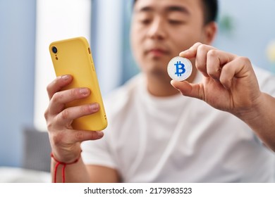 Young Chinese Man Using Smartphone Holding Bitcoin At Home