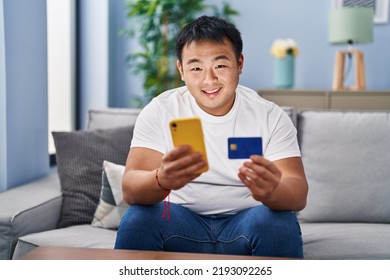 Young Chinese Man Using Smartphone And Credit Card Sitting On Sofa At Home