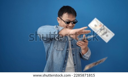Young chinese man wearing thug life glasses throwing dollars over isolated blue background