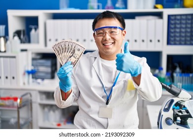 Young Chinese Man Working At Scientist Laboratory Holding Money Smiling Happy And Positive, Thumb Up Doing Excellent And Approval Sign 