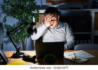 Young Chinese Man Working Using Computer Laptop At Night Peeking In Shock Covering Face And Eyes With Hand, Looking Through Fingers With Embarrassed Expression. 