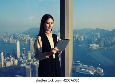 Young Chinese Successful Woman CEO Is Reading Financial News In Internet Via Portable Digital Tablet, While Is Standing In Modern Office Interior Against Window With View Developed New York Cityscape