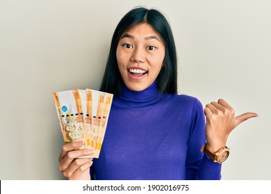 Young chinese woman holding 500 philippine peso banknotes pointing thumb up to the side smiling happy with open mouth 