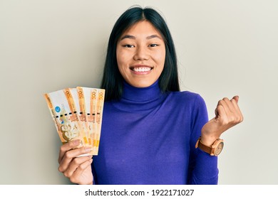 Young chinese woman holding 500 philippine peso banknotes screaming proud, celebrating victory and success very excited with raised arm 