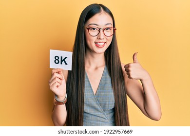 Young chinese woman holding 8k resolution technology paper smiling happy and positive, thumb up doing excellent and approval sign 