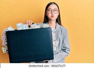 Young chinese woman holding briefcase full of dollars puffing cheeks with funny face. mouth inflated with air, catching air. 