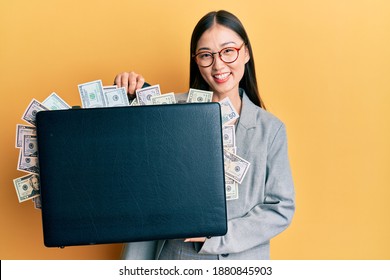 Young chinese woman holding briefcase full of dollars smiling and laughing hard out loud because funny crazy joke. 