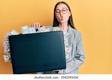Young chinese woman holding briefcase full of dollars making fish face with mouth and squinting eyes, crazy and comical. 