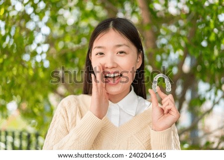 Young Chinese woman holding invisible braces at outdoors shouting with mouth wide open