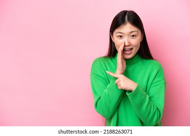 Young Chinese woman isolated on pink background pointing to the side to present a product and whispering something