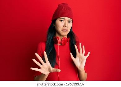 Young chinese woman listening to music using headphones disgusted expression, displeased and fearful doing disgust face because aversion reaction. with hands raised 