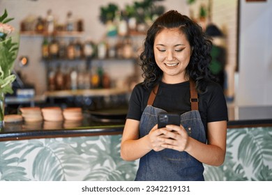 Young chinese woman waitress smiling confident using smartphone at restaurant