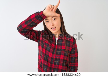 Young chinese woman wearing casual jacket standing over isolated white background making fun of people with fingers on forehead doing loser gesture mocking and insulting.