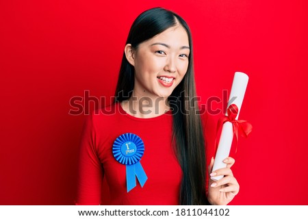 Young chinese woman wearing first place badge holding diploma looking positive and happy standing and smiling with a confident smile showing teeth 