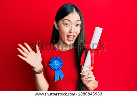 Young chinese woman wearing first place badge holding diploma celebrating achievement with happy smile and winner expression with raised hand 