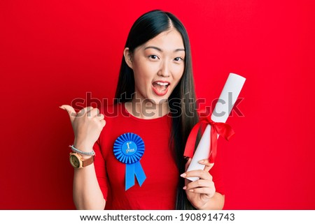 Young chinese woman wearing first place badge holding diploma pointing thumb up to the side smiling happy with open mouth 