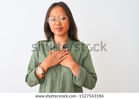 Young chinese woman wearing green shirt and glasses over isolated white background smiling with hands on chest with closed eyes and grateful gesture on face. Health concept.