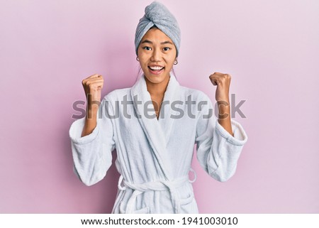 Young chinese woman wearing shower towel cap and bathrobe celebrating surprised and amazed for success with arms raised and open eyes. winner concept. 