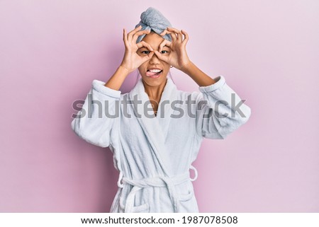 Young chinese woman wearing shower towel cap and bathrobe doing ok gesture like binoculars sticking tongue out, eyes looking through fingers. crazy expression. 