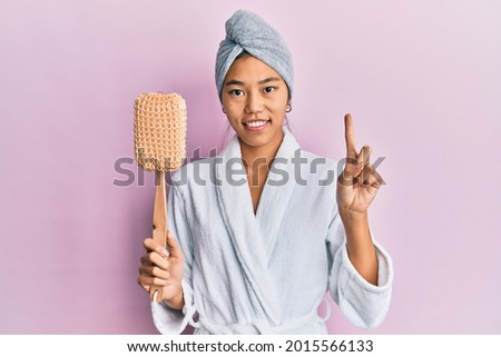 Young chinese woman wearing shower towel cap and bathrobe holding sponge smiling with an idea or question pointing finger with happy face, number one 