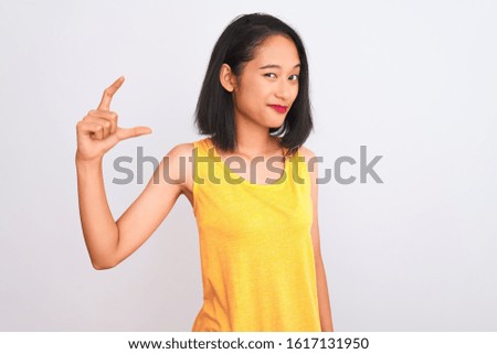 Young chinese woman wearing yellow casual t-shirt standing over isolated white background smiling and confident gesturing with hand doing small size sign with fingers looking and the camera. Measure 
