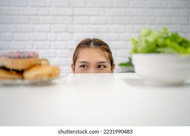 Young chubby asian woman standing hiding under table with fresh vegetables looking at plate with donuts and sweets. - Powered by Shutterstock