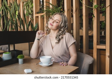 A Young Chubby Blonde Woman In A Beige Blouse Drinks Coffee In A Cafe Sits Near A Large Panoramic Window. Thoughtful Girl With A Cup Of Tea In Light Modern Wooden Interior