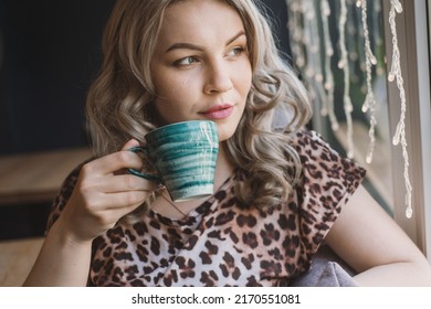 A Young Chubby Blonde Woman In A Leopard Blouse Drinks Coffee From In A Cafe Sits Near A Large Panoramic Window. Portrait Of Pretty Plus Size Girl