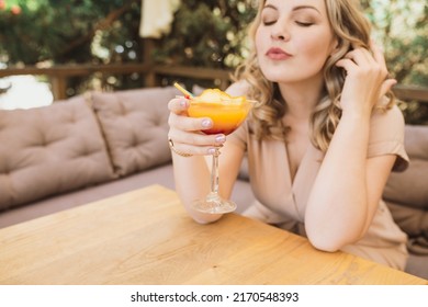 Young Chubby Pretty Blonde Woman With Curls Is Drinking A Summer Cocktail In Orange Color On The Terrace In A Cafe. Selective Focus And Glass Close Up