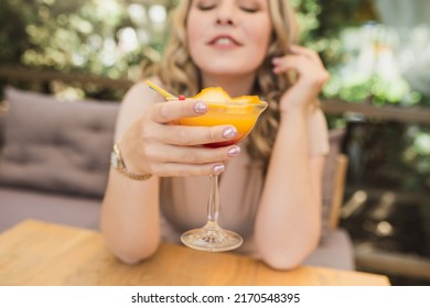 Young Chubby Pretty Blonde Woman With Curls Is Drinking A Summer Cocktail In Orange Color On The Terrace In A Cafe.  Selective Focus And Glass Close Up