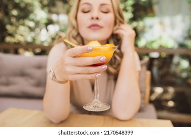 Young Chubby Pretty Blonde Woman With Curls Is Drinking A Summer Cocktail In Orange Color On The Terrace In A Cafe. Selective Focus And Glass Close Up