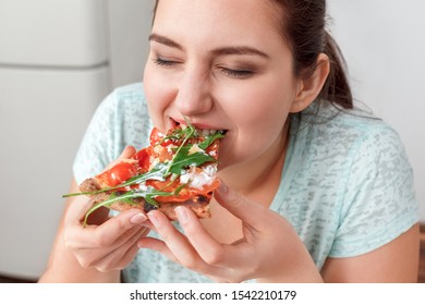 Young Chubby Woman Sitting At Table In Kitchen Binge Eating Eating Slice Of Pizza Close-up Closed Eyes Smiling Delightful