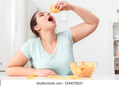Young chubby woman sitting at table in kitchen breaking diet eating potato chip mouth opned for new portion hungry