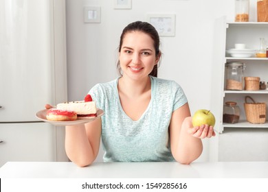 Young Chubby Woman Sitting At Table In Kitchen Choosing Between Plate With Doughnut And Cake And Healthy Apple Looking Camera Smiling Positive