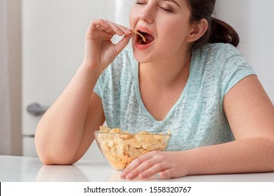 Young chubby woman sitting at table in kitchen breaking diet eating potato chip closed eyes smiling delightful close-up