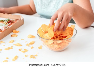 Young chubby woman sitting at table in kitchen with box of pizza breaking diet eating potato chip from bowl close-up