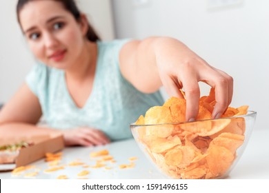 Young chubby woman sitting at table in kitchen breaking diet eating potato chip taking portion excited close-up blurred background