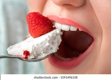 Young chubby woman sitting at table in kitchen holding teaspoon eating cake with raspberry breaking diet open mouth close-up