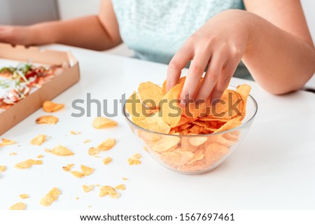 Young chubby woman sitting at table in kitchen with box of pizza breaking diet eating potato chip from bowl close-up
