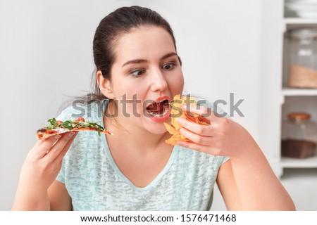 Young chubby woman sitting at table in kitchen binge eating eating slice of pizza and chips hungry close-up