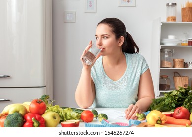 Young Chubby Woman Standing In Kitchen Sitting At Table Holding Glass Drinking Pure Water Looking Out The Window Pensive