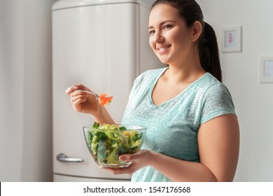 Young Chubby Woman Standing In Kitchen Eating Healthy Fresh Green Salad Looking Aside Smiling Cheerful