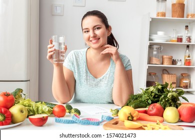 Young Chubby Woman Standing In Kitchen Sitting At Table Holding Glass Drinking Pure Water Looking Camera Smiling Cheerful