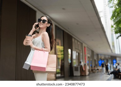 A young classy asian woman in a light top and khaki pants and wearing stylish sunglasses walking around an al fresco mall carrying shopping bags and talking over the phone. - Powered by Shutterstock