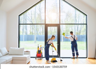 Young Cleaners In Blue Uniform Mopping Wooden Floor, Cleaning Big Panoramic Window, Preparing Country House For Selling. Cleaning Service Concept