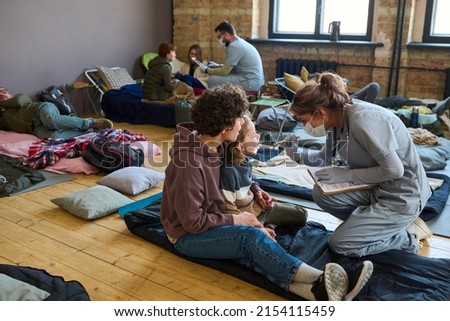 Young clinician with document checking up throat of little boy sitting on sleeping place on the floor of refugee camp next next to his mother