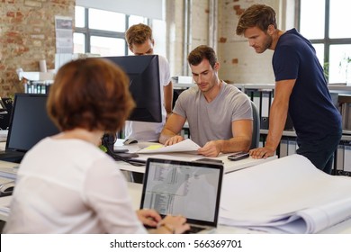 Young colleagues working together in office gathered at desk with computers and papers, with woman with laptop blurred in foreground