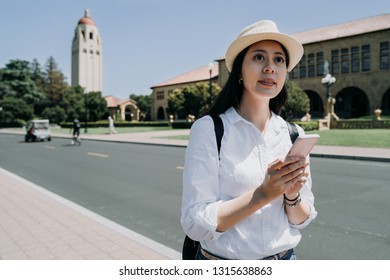 Young College Girl Join Short Term Study Tour Program In University In Usa. Female Student Freshman Relaxing Sightseeing Holding Cellphone Looking Online Guide Book In Stanford On Sunny Day Wear Hat.