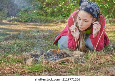 Young College Girl Wearing Mountain Clothes Blows To Burn Some Pine Needles And Start A Small Campfire During A Hiking Day.