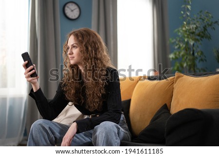 A young college girl is wondering what to write back to an email she received. The girl is sitting on couch in a cozy living room using cell phone. The smiling teenage girl is studying remotely.